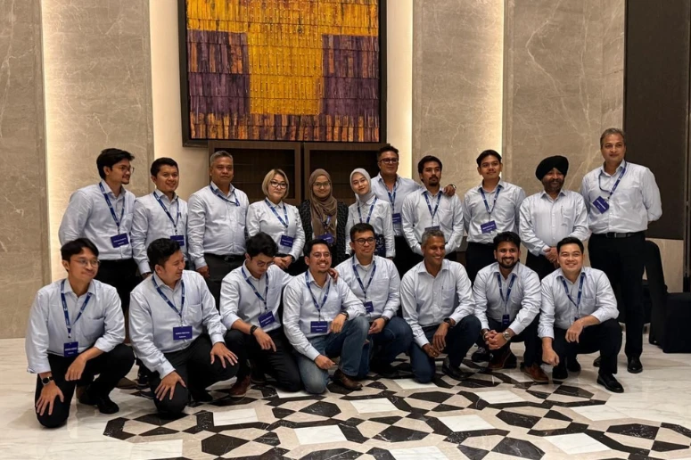 A group photo of the Forbes Marshall Indonesia team posing in a modern building lobby with marble walls and a geometric patterned floor. The team consists of 18 professionals—13 men and 5 women—all dressed in light blue button-down shirts and dark trousers, with several wearing blue company lanyards. The group is arranged in two rows: the front row is kneeling or crouching, while the back row stands in front of a large, textured yellow and purple abstract art piece.