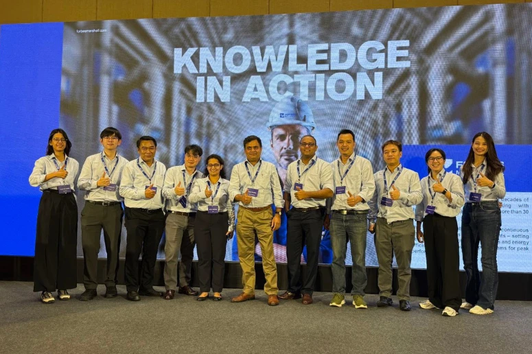 A group of 11 Forbes Marshall Thailand team members—7 men and 4 women—standing in a row on a stage. They are positioned in front of a large digital screen displaying a technician in a hard hat and the bold text "KNOWLEDGE IN ACTION". Most team members are wearing light blue button-down shirts with blue lanyards and giving a "thumbs up" gesture. The Forbes Marshall logo is visible on the right side of the screen.
